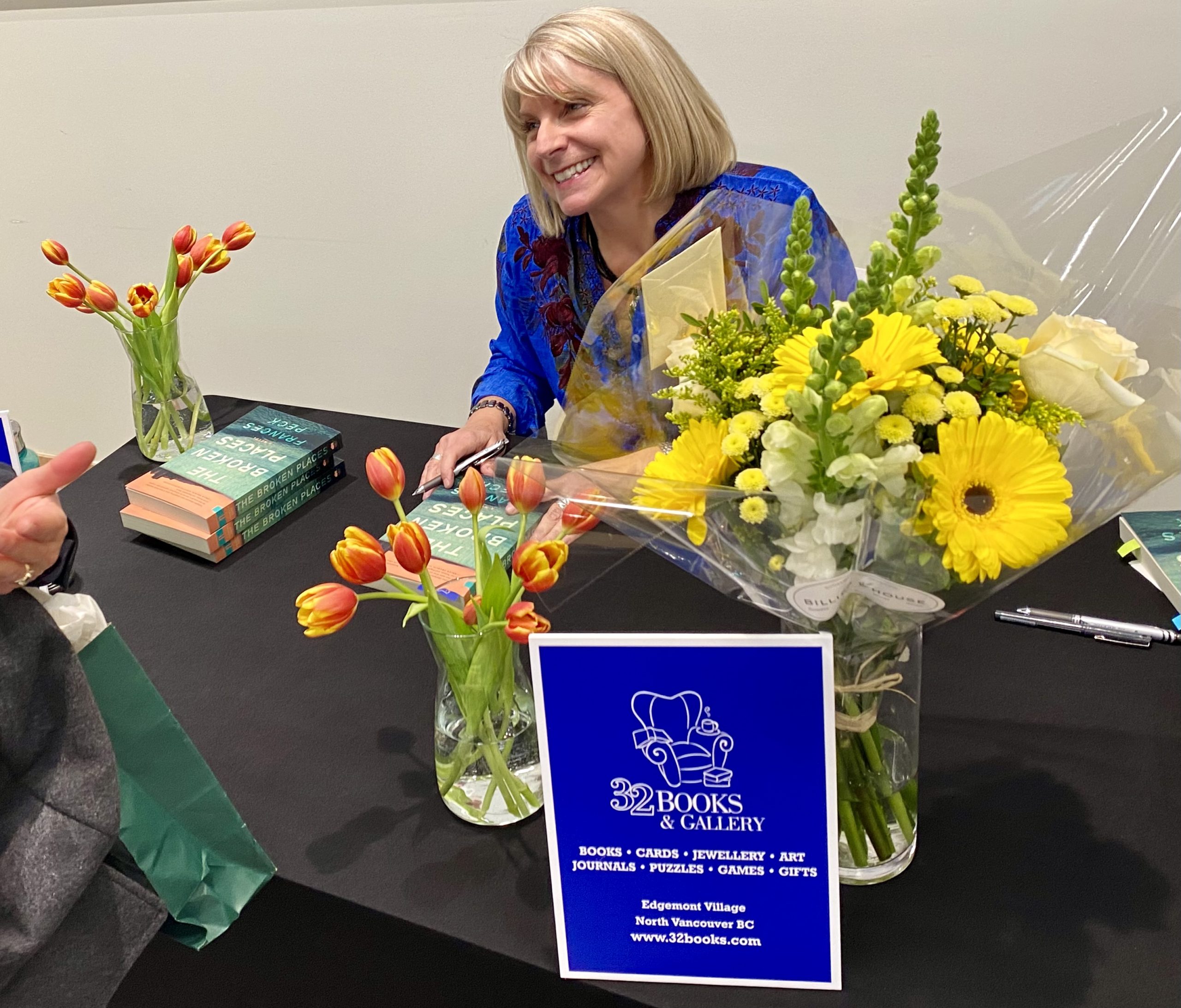 Author Frances Peck sitting at a table, surrounded by flowers, signing copies of her novel, The Broken Places.