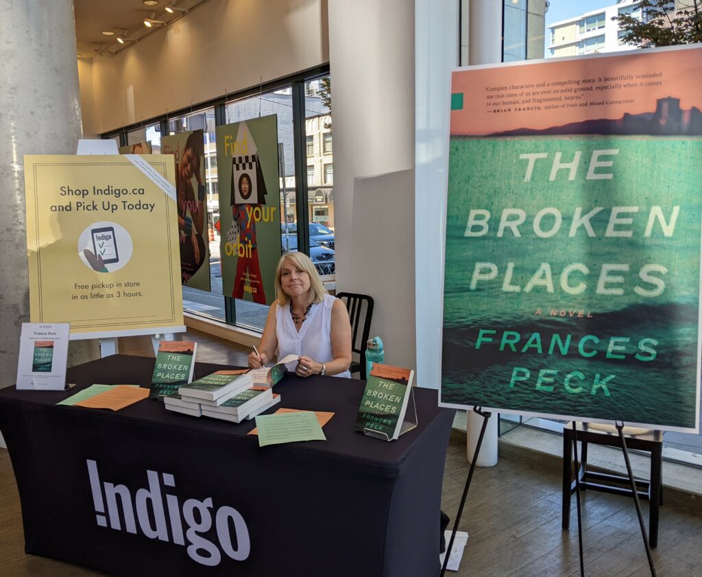 Photo of Frances Peck sitting at Indigo table, ready to sign books, which are set up in front of her.