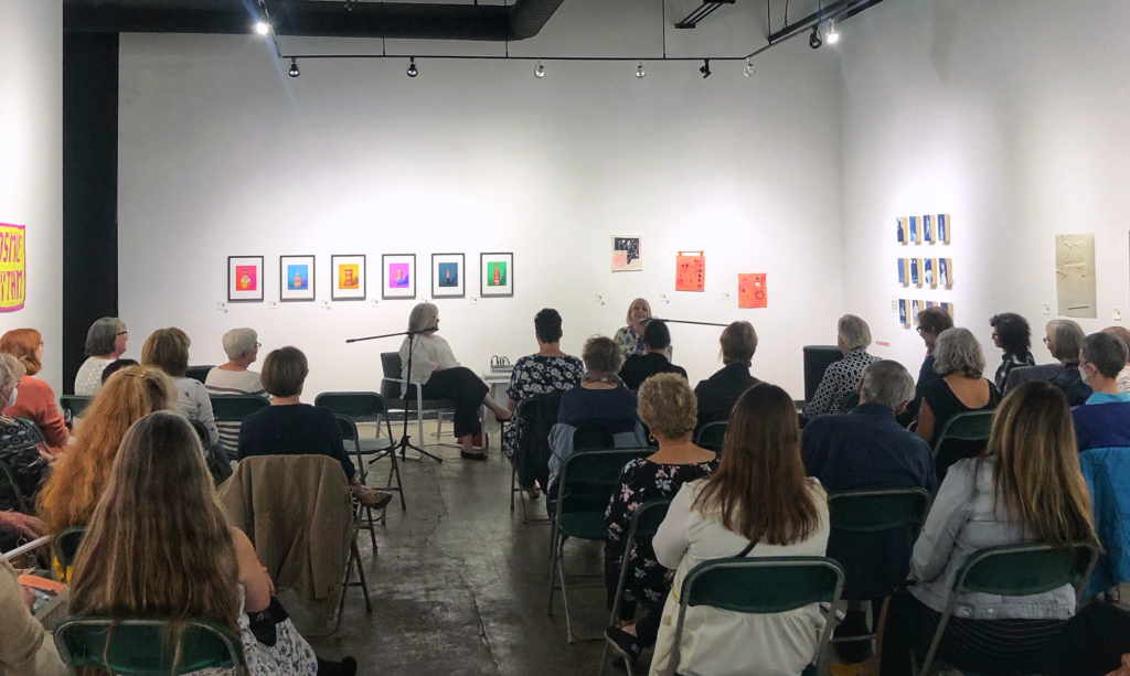 Photo taken from back of the room at Glass Bookshop, showing the backs of attendees, and emcee Virginia Durksen and Frances Peck seated at mics at the front.