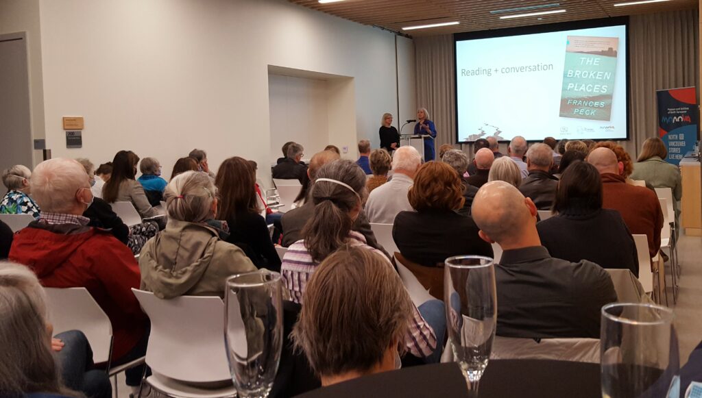 Photo from back of the room, showing backs of attendees, with emcee Eve Lazarus and Frances Peck at the podium up front. A slide on the screen behind them shows the cover of The Broken Places and says "Reading + Conversation."