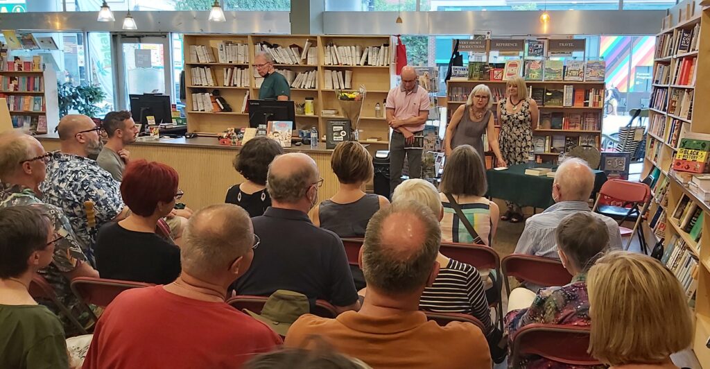 Photo taken over the heads of attendees, looking toward the front of the room at Perfect Books, where authors Norm Boucher and Frances Peck stand with emcee Moira White.