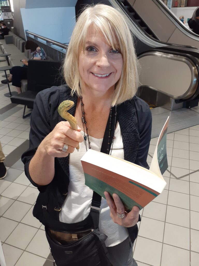Frances Peck holding a cobra-shaped pen, about to sign a copy of her novel, The Broken Places.