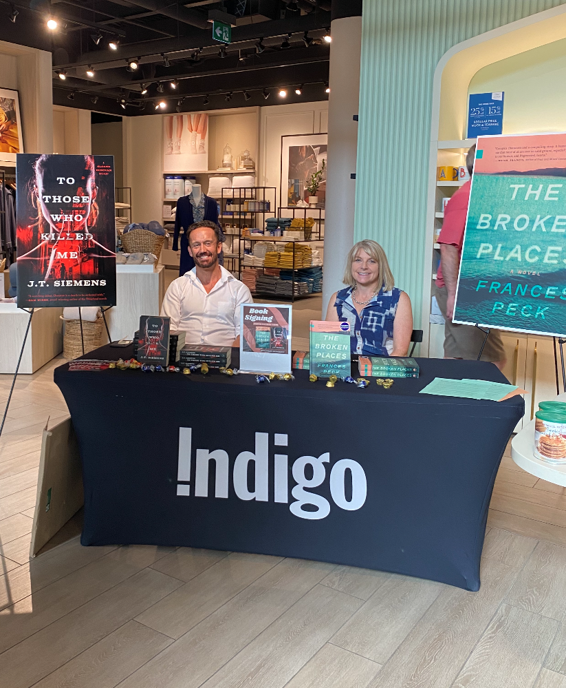 Photo of J.T. Siemens on the left and Frances Peck on the right, sitting side by side at an Indigo table, ready to sign books, which are set up in front of them. Behind them are large posters of their book covers.