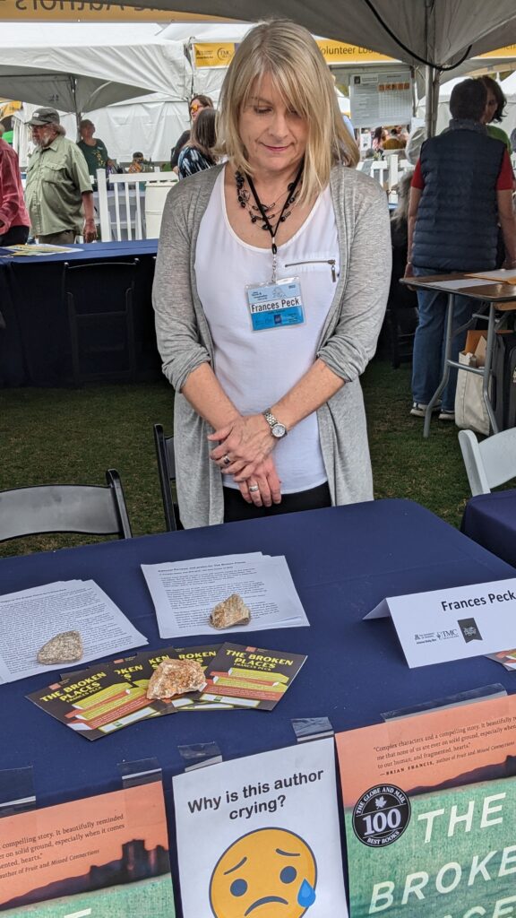 Frances Peck standing under a large tent and ehind her book sales table. On the table are piles of leaflets and handouts. Hanging from the front of the table are posters of the cover of The Broken Places, along with a poster of a crying-face emoji plus the caption "Why is this author crying?"
