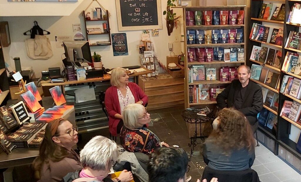 Indoors at Windowseat Books. At the front, seated with their backs to the sales counter, are Frances (left) and Danial (right). The heads of five audience members are in the foreground. To the left is a table piled with copies of The Sum of One Man's Pleasure and Uncontrolled Flight.