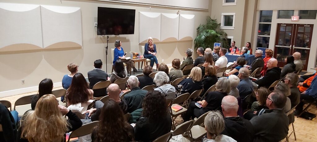 Shot from the back of the room: group of people sitting and facing platform where Susan Safyan (left) and Frances Peck (right) are seated and in conversation.