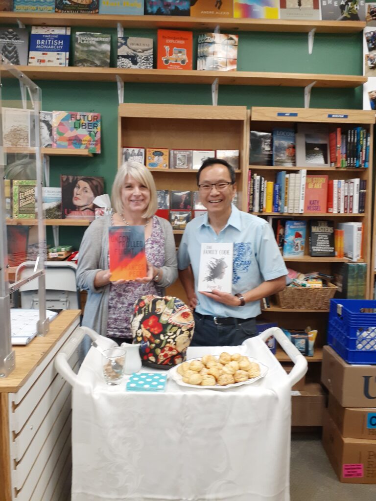 Frances (left) and Wayne Ng (right), both holding copies of their novels, standing in front of bookstore shelves. In front of them is a cloth-covered trolley that is set with a teapot, napkins, and a plate of scones.
