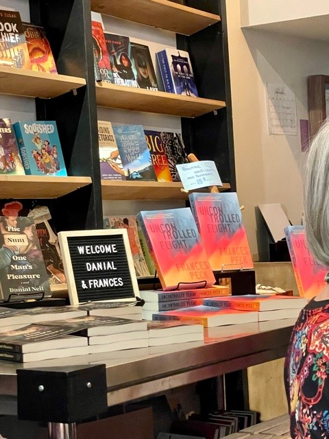 Display table set up in front of bookstore bookshelves. On the table are copies of The Sum of One Man's Pleasure and Uncontrolled Flight, with a sign between the two that says "Welcome Danial and Frances