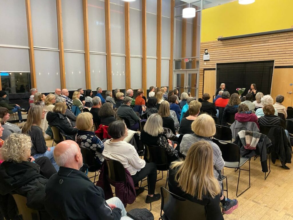 Photo of Frances Peck and Tara McGuire sitting at the front of a large windowed room, taken from the back and showing the rows of attendees in between.