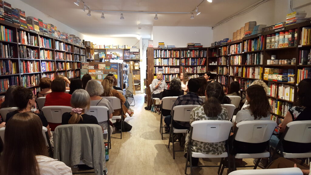 Photo from the back of the room at Iron Dog Books, showing the backs of attendees' heads and the three authors seated at the front. Left to right, Frances (speaking into the mic), Robin Yeatman, and Lisa Brideau