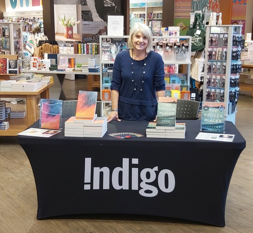 Frances in a bookstore, standing behind a table with an Indigo cloth on it. On the table are piles of the book Uncontrolled Flight and The Broken Places, as well as bookmarks and sheets of reviews.