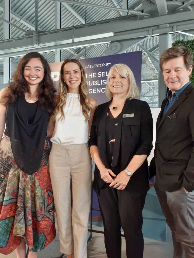 Four people stand side by side: from left to right, a young white woman with long brown hair, wearing s black top and colourful skirt; a young white woman with long dirty blond hair, wearing a sleeveless white blouse and beige pants; an older white woman with shoulder-length blond hair wearing black pants, top and jacket; and an older white man with short brown hair wearing a brown suit jacket and lighter pants.